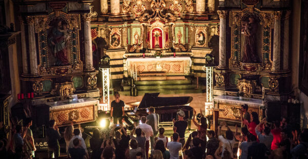 A pianist performing in front of an audience inside a baroque-style church, with the crowd standing and applauding, and intricate church decorations and altar in the background.