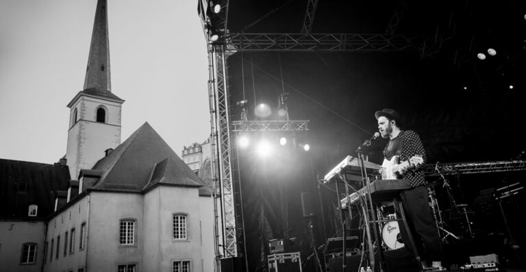 A black and white photo of a musician playing a keyboard and singing into a microphone on an outdoor stage with concert lighting, a church spire, and building in the background.