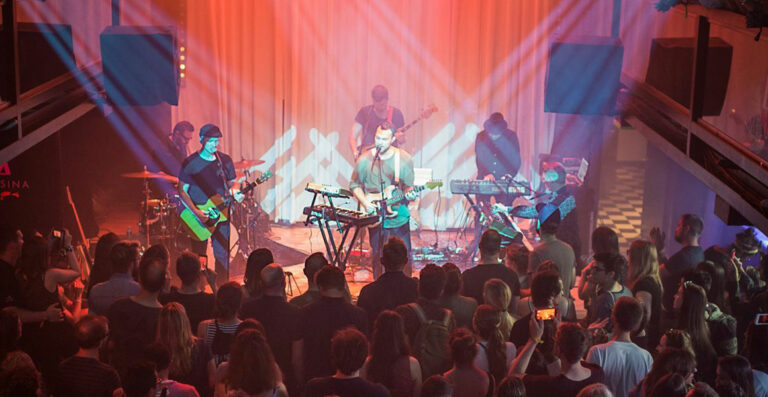A band performs on stage in a dimly lit venue with colorful stage lighting, in front of an audience of spectators.