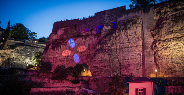 A large, illuminated rock wall with colored light projections, flanked by greenery and architectural structures at dusk.