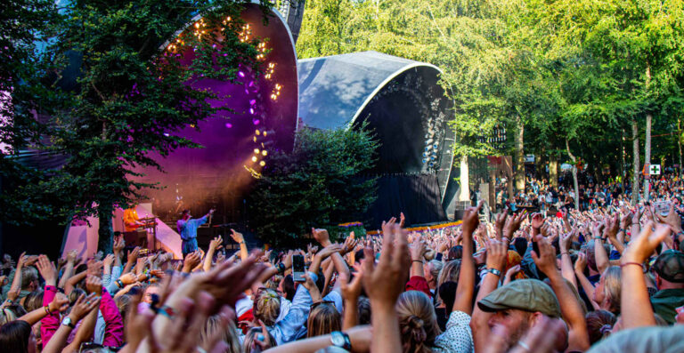 Outdoor music festival scene with a crowd of people raising their hands towards a stage with bright lights and trees in the background.