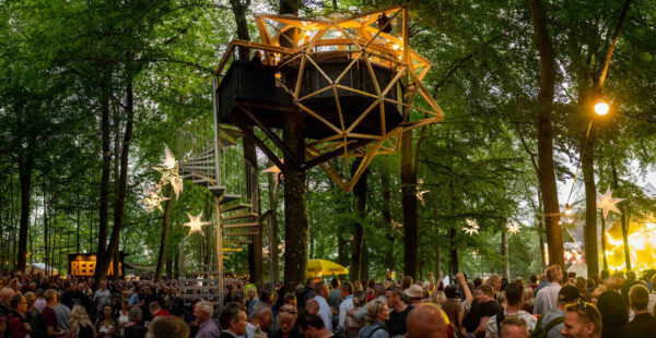 A crowd of people gathered at an outdoor event in a forested area with a unique treehouse structure illuminated from within.
