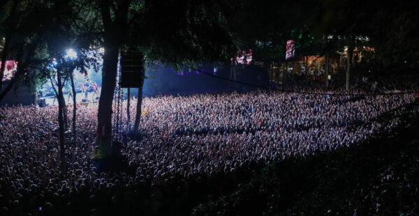A large crowd of people at an outdoor nighttime concert with bright stage lights and large screens among trees.