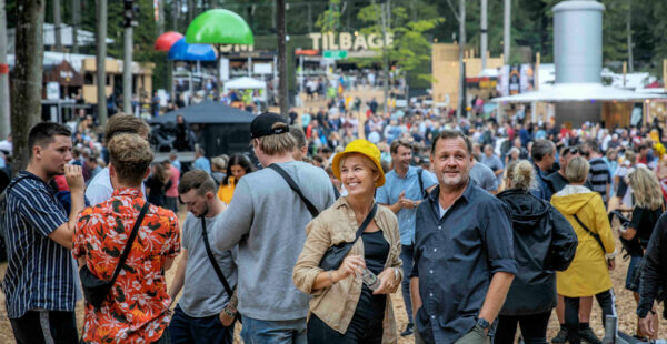A crowd of people at an outdoor festival, with some individuals standing out in the foreground, and a sign reading 'TILBAGE' in the background.