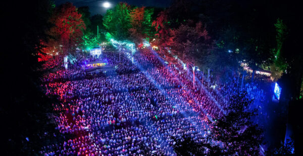 Nighttime aerial view of an outdoor music festival with large crowds illuminated by colorful stage lights, surrounded by trees.
