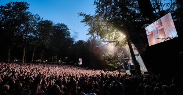 A large outdoor concert with a cheering crowd, stage lights, a big screen displaying a performer, and surrounded by trees during dusk.