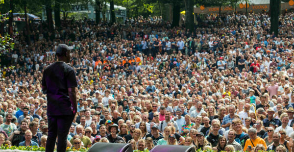 A performer stands on stage facing a large outdoor crowd of spectators under a sunny sky.