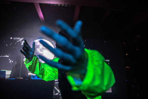A person with an out-of-focus hand reaching toward the camera, wearing a neon green jacket, with a backdrop of dimly lit stage and visual effects.
