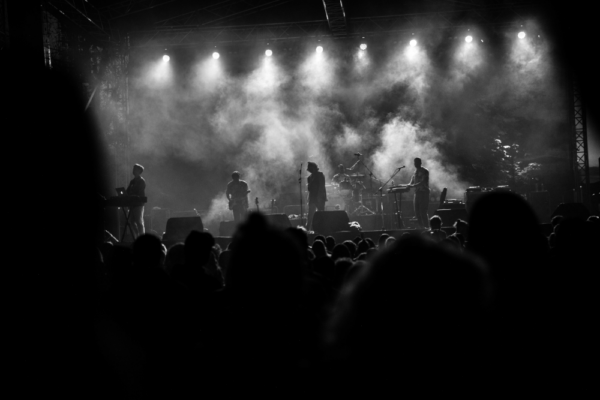 A black and white photo of a concert showing a band performing on stage with backlighting, silhouetting the musicians, and an audience in the foreground.