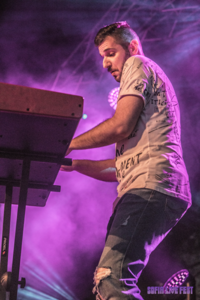 A focused male musician playing an electronic keyboard on stage with purple stage lighting in the background. He's wearing a printed t-shirt and ripped jeans.
