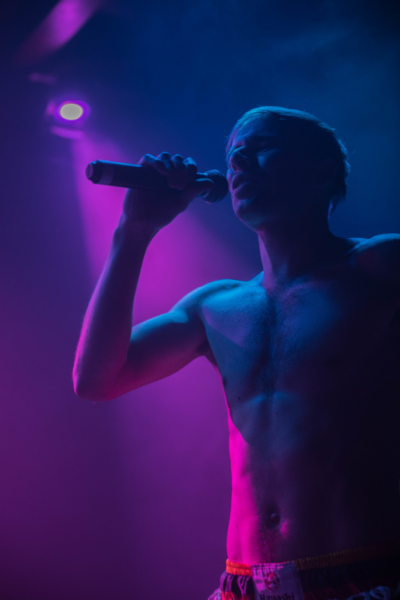 A shirtless male performer singing into a microphone onstage with purple and pink stage lighting.
