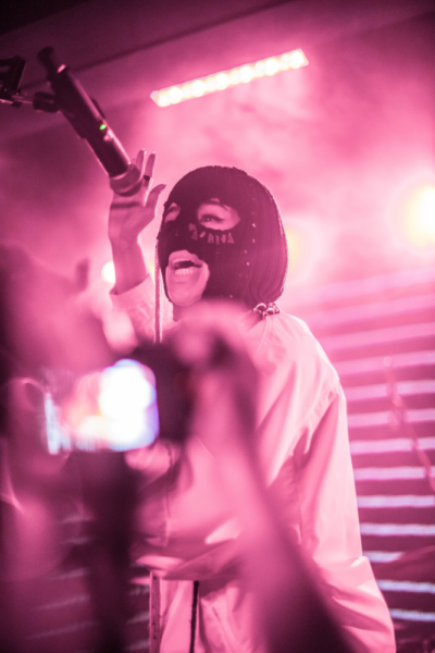 A performer in a balaclava and white outfit singing into a microphone on a stage with pink lighting and motion blur effects.