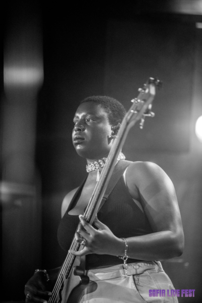 A black-and-white photo of a focused woman playing an electric bass guitar onstage, with a blurred backdrop featuring the text 