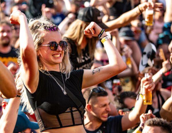 A woman with sunglasses and braided hair enjoying a music festival, smiling and raising her arms in the air among a crowd of people, some holding beers.