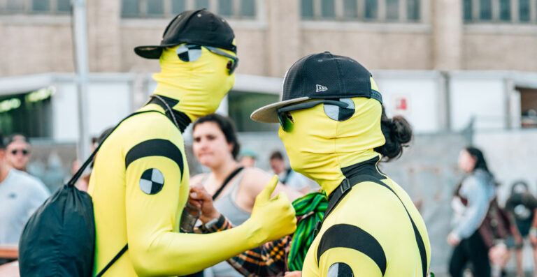 Two people in yellow and black full body suits and sunglasses, with one wearing a black cap, are seen from the back in an outdoor, crowded setting.