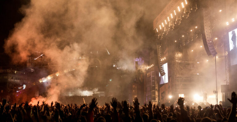 A crowded outdoor music concert at night with the stage enveloped in smoke, spotlights, and the silhouettes of raised hands in the audience.