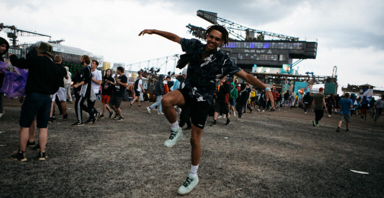 A person with a playful pose dancing at a festival with other attendees and festival structures in the background.