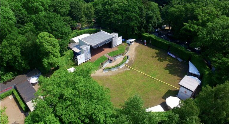 Aerial view of an outdoor amphitheater with seating and stage surrounded by green trees in a park setting.