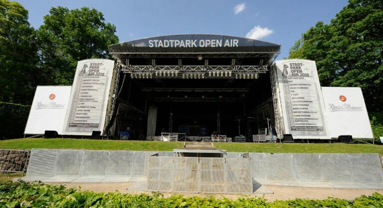 An empty outdoor stage at Stadtpark Open Air with event banners on either side, metal barriers in the foreground, and trees in the background under a clear sky.