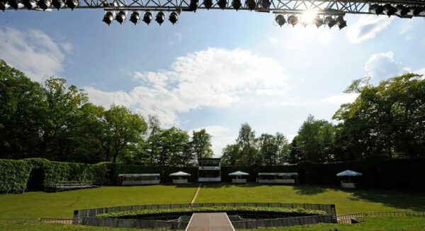Outdoor stage with lighting rig, empty seating area, and green trees under a blue sky with white clouds.