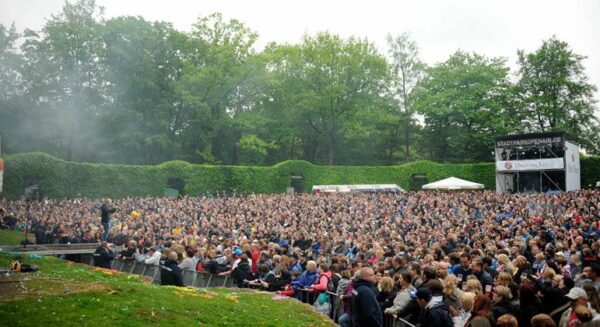 A large crowd of spectators at an outdoor concert with lush green trees surrounding the area and a stage in the background.