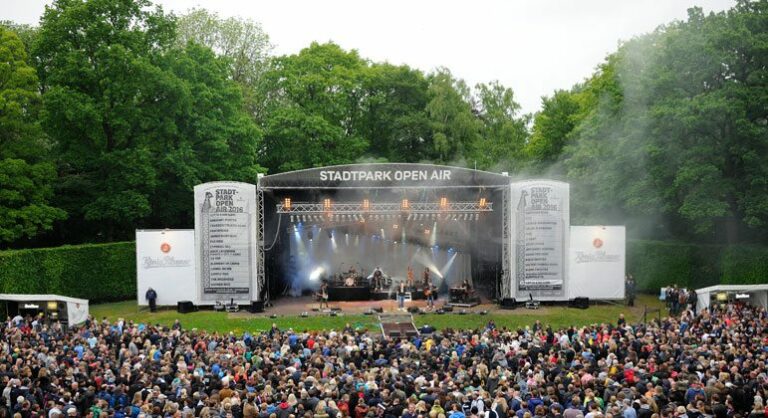 A large crowd of people gathered for an outdoor concert at Stadtpark Open Air with a band performing on stage and trees surrounding the venue.