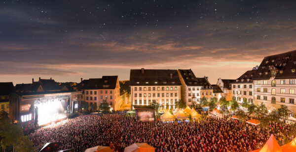 An outdoor concert at night with a large crowd gathered in front of a stage, surrounded by historic buildings, against a backdrop of a starry sky.