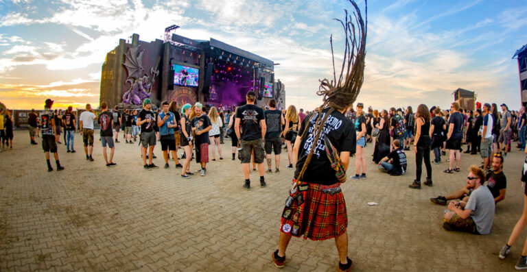 The image shows a vibrant outdoor music festival at sunset with a crowd of people, some standing and others sitting on the ground. In the foreground, a person with long dreadlocks headbanging to the music can be seen. The stage in the background features a massive screen displaying the performers, and elaborate decorations including a large dragon sculpture. The attendees are dressed in various casual and alternative fashions, indicative of the metal music scene.