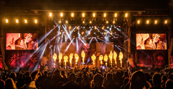 Large crowd at an outdoor rock concert with intense stage lighting and pyrotechnics, large screens displaying performers, and a dynamic, energetic atmosphere.