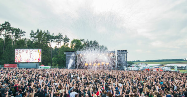 A large outdoor music festival crowd with many fans raising their hands towards a stage with bright lights and pyrotechnics, set against a backdrop of trees and a clear sky.