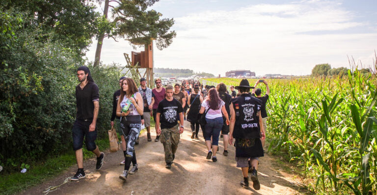 A group of people walking down a dirt path flanked by trees and a cornfield, some dressed in alternative fashion, with a clear sky above.