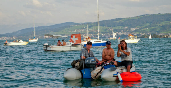 Three men on a small inflatable boat on a lake with other boats and hills in the background.