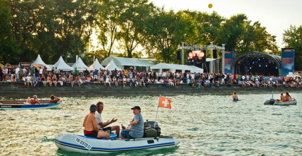 An outdoor festival by a body of water, with a crowd of people on the shore and some in boats, including one flying a Swiss flag. A stage with screens is visible in the background.