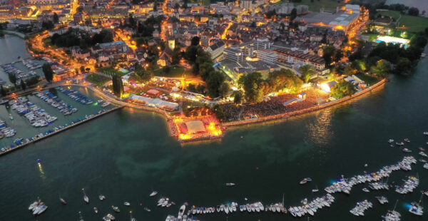 Aerial view of an evening outdoor event on a peninsula with a large crowd, illuminated stages, food stands, boats on the surrounding waters, and adjacent city lights.