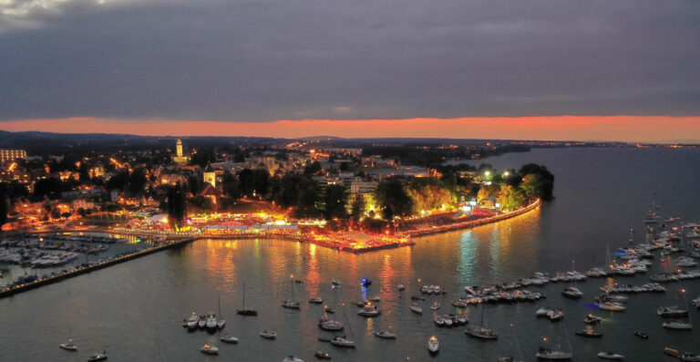 Aerial view of a lakeside city at dusk, with illuminated waterfront and boats in the water, against an orange and blue sky.