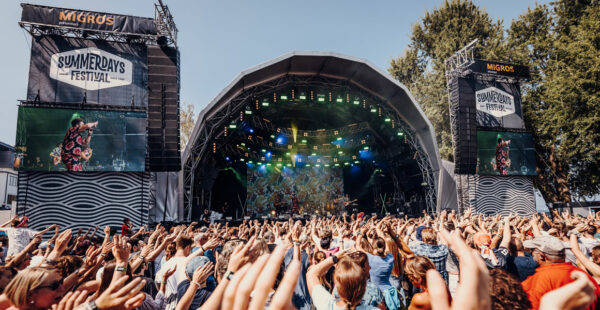 A crowd of festival-goers with their hands up, enjoying a live performance at an outdoor stage during the Summerdays Festival, with large screens on either side of the stage showing a musician.