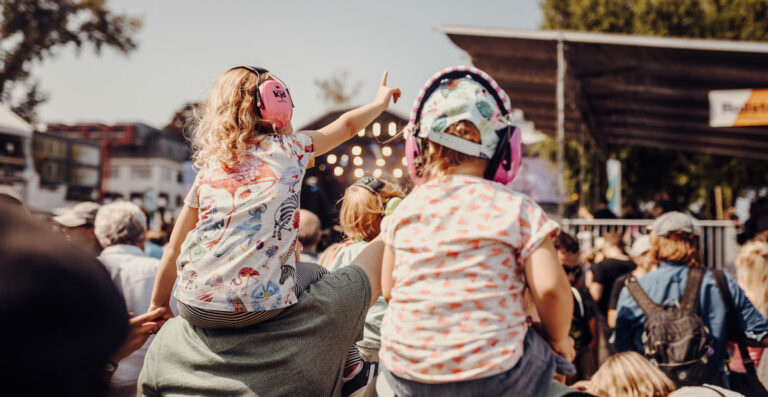 Two children wearing protective headphones are sitting on adults' shoulders at an outdoor event, with the focus on the child on the left pointing ahead enthusiastically.