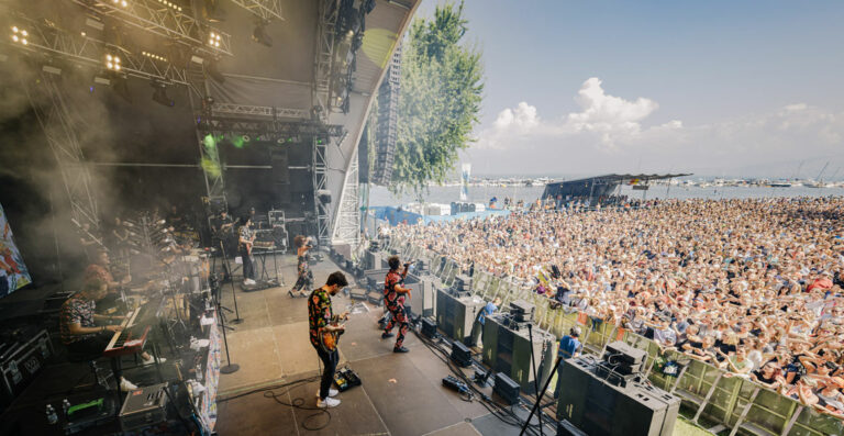 A wide-angle view of a lively outdoor music festival showing band members performing on stage and a large, enthusiastic crowd enjoying the event, with a clear sky above and trees and a body of water in the background.