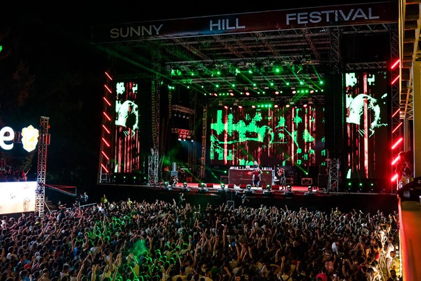 A lively outdoor music festival at night with a large crowd of people and hands raised in the air, in front of a brightly lit stage with large screens displaying visual effects and the text 