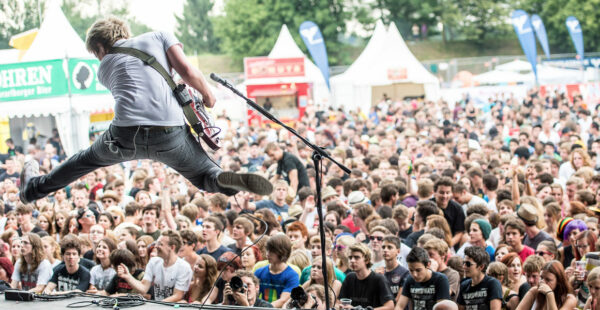 A guitarist jumping in mid-air during a live performance in front of a densely packed audience at an outdoor music festival.
