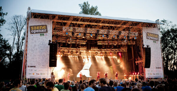 Outdoor music festival scene with a large stage and a performance in progress, surrounded by a crowd of spectators in the evening. Banners with 'Szene Openair' are displayed on either side of the stage.
