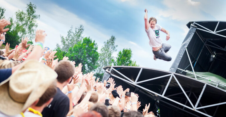A person is stage diving into a crowd at an outdoor concert, with outstretched arms and uplifted faces of audience members indicating excitement and anticipation.