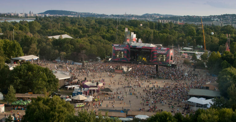 Aerial view of a crowded outdoor music festival with large stage and screens, surrounded by trees, with a city skyline in the background during daylight.