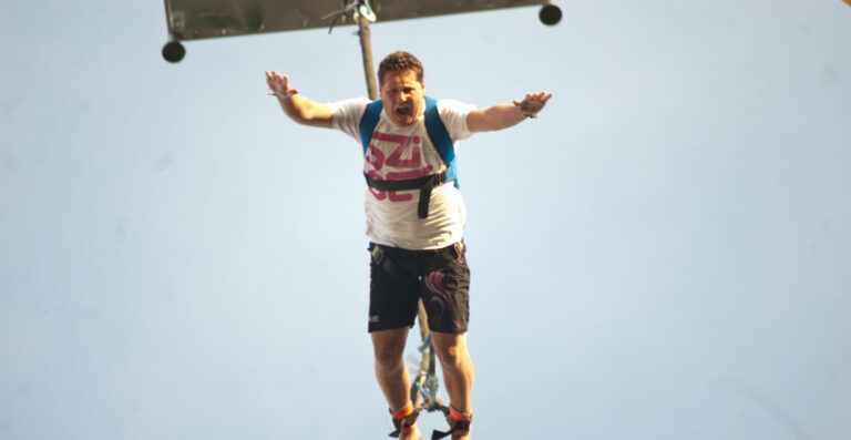 A person in a harness appears to be bungee jumping against a clear blue sky, with their arms outstretched and a slight look of apprehension on their face.