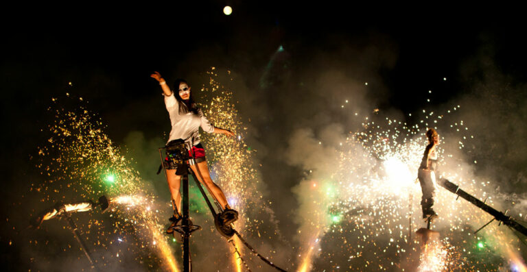 A performer on stilts wearing a mask participates in a nighttime event, surrounded by a vibrant display of fireworks and smoke.