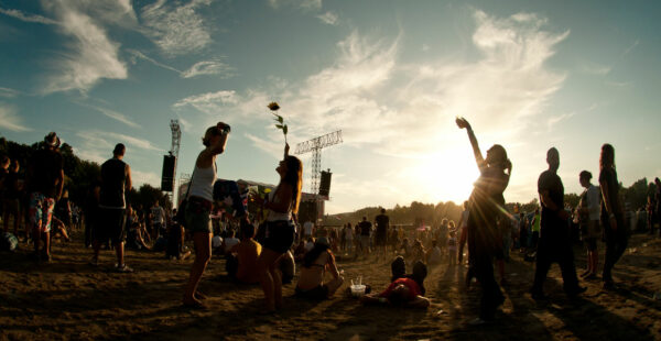 Outdoor music festival scene with people standing and sitting on the grass, enjoying the event with a stage in the background and the sun low on the horizon, casting a warm backlight.