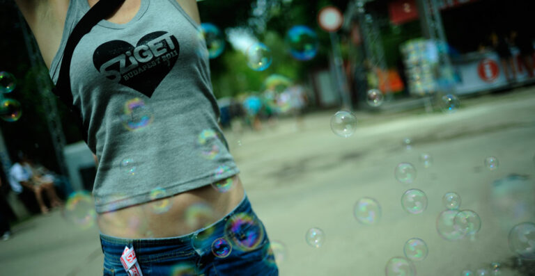 A close-up, selective focus image of soap bubbles floating in the air with the partial torso of a person wearing a Sziget Budapest 2011 t-shirt in the background.