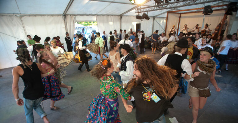 A lively folk dance event inside a tent with people in various traditional and casual outfits dancing while musicians with violins and a double bass perform on stage in the background.