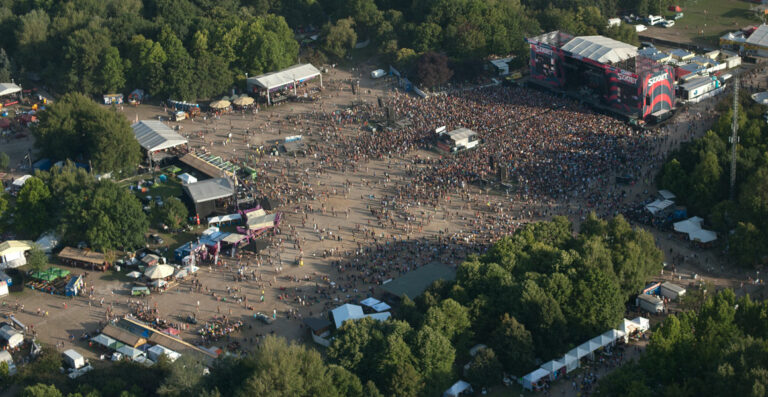 Aerial view of an outdoor music festival with large crowds gathered around the main stage, vendor booths, and open areas surrounded by trees.