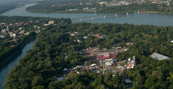 Aerial view of a large outdoor music festival in a green park area with multiple stages and a dense crowd of attendees, surrounded by a river and a bridge in the background.